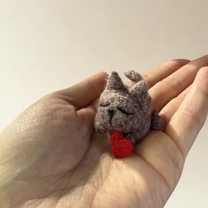 Könnte beinhalten: Eine kleine, nadelgefilzte graue Katzenfigur mit spitzen Ohren und geschlossenen Augen, die ein winziges, leuchtend rotes Herz zwischen ihren Vorderpfoten hält. Die strukturierte, flauschige Wollskulptur zeigt sichtbare Faserdetails.