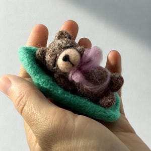 Puede incluir: Un oso de peluche marrón afieltrado hecho a mano con un lazo rosa, descansando sobre una pequeña estera verde de fieltro. El oso tiene un hocico marrón claro y está acostado. El artículo se sostiene en una mano.