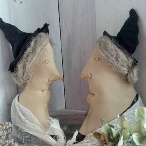 May include: Two whimsical fabric dolls with long noses and gray hair, wearing black witch hats and draped in gauze. The dolls are positioned facing each other, creating a unique and artistic display. Decorative flowers and lace add to the vintage aesthetic.