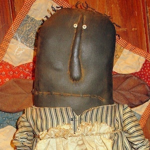 May include: A handmade rag doll with a brown face, white eyes, and a brown nose. The doll is wearing a striped dress with a white ruffle collar and brown fabric wings.