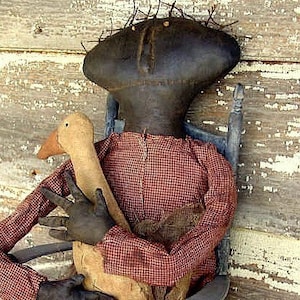 May include: A rag doll with a black head and red and white checkered shirt is holding a tan goose. The doll is sitting in a wooden chair.