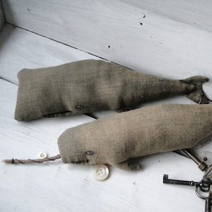 May include: Two gray fabric whale-shaped pillows with button eyes and a twig attached to one of the whales. The whales are on a white wooden surface with old keys and a button.