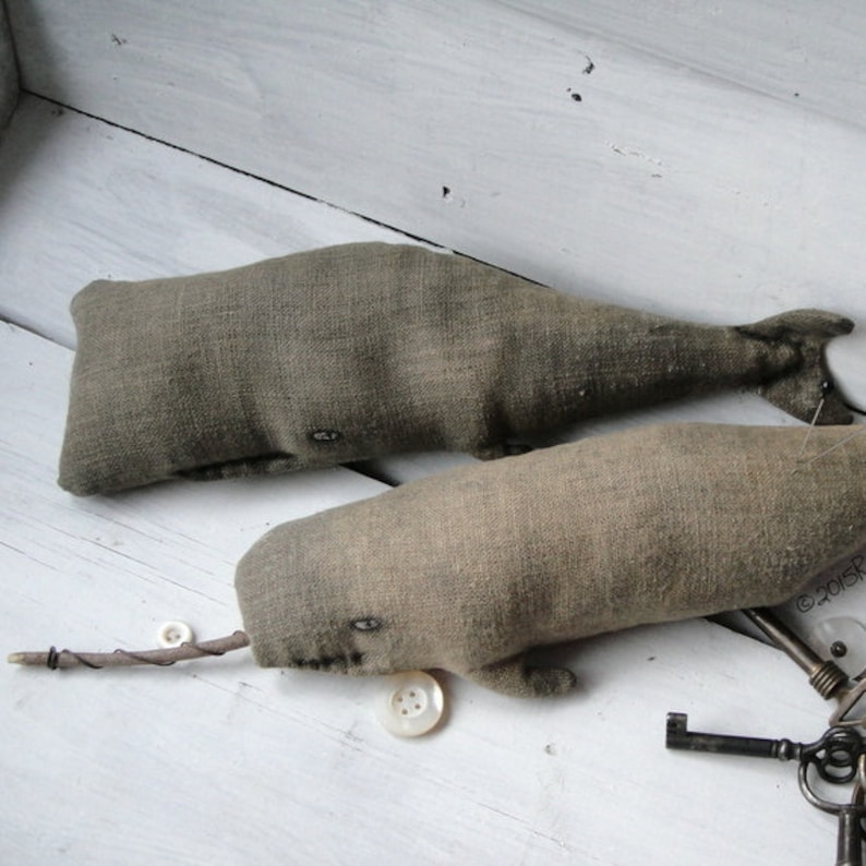 May include: Two gray fabric whale-shaped pillows with button eyes and a twig attached to one of the whales. The whales are on a white wooden surface with old keys and a button.