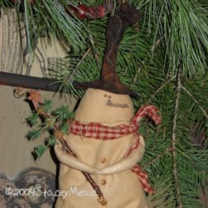 May include: A handmade snowman figurine with a brown felt hat, a red and white plaid scarf, and a twig in its hand. The snowman is made of beige fabric and has a painted face with black eyes and a brown smile.