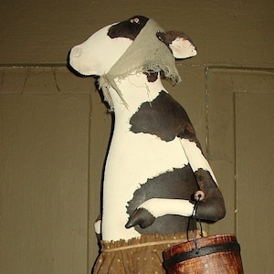 May include: A black and white cow figurine with a brown fabric skirt and a brown bucket. The cow is wearing a brown fabric head scarf.