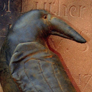 May include: A close-up of a dark gray crow figurine with a long, pointed beak. The crow is perched on a textured, brown surface.