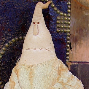 May include: A white fabric ghost doll with a stitched-on mouth and eyes. The doll is wearing a white fabric robe and is sitting against a dark brown background.