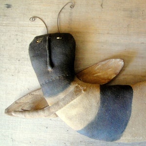 May include: A handmade black and tan bee with wire antennae and a stitched face. The bee is lying on a wooden surface.