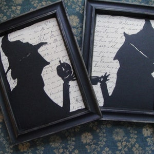 May include: Two black framed shadow boxes with white paper backgrounds and black paper cutouts of witches holding pumpkins. The text in the background is handwritten in black ink.