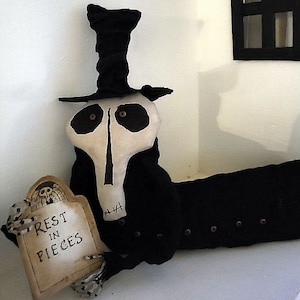 May include: A black and white skeleton doll wearing a black top hat and a black coat. The doll is holding a sign that says "Rest in Pieces".