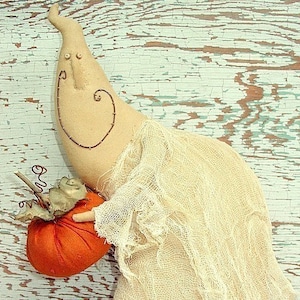 May include: A white fabric ghost doll with a stitched-on face and a large orange pumpkin in its hand. The doll is sitting on a light blue wooden surface.