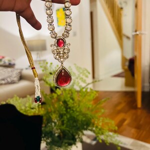 Puede incluir: Un collar de oro con un colgante en forma de lágrima que presenta una gran piedra roja. El collar está adornado con piedras blancas más pequeñas.