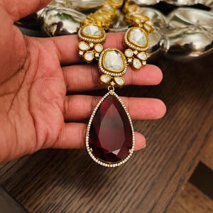 Pode incluir: Um colar de ouro com um grande pingente vermelho em forma de lágrima. O pingente é rodeado por diamantes menores. O colar é feito de ouro e tem um design tradicional indiano.