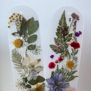 May include: Two clear bookmarks featuring pressed flowers and leaves. The bookmark on the left has white, yellow, and green flowers and leaves. The bookmark on the right has purple, pink, blue, and green flowers and leaves.