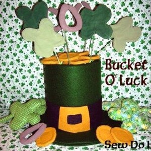 Puede incluir: Un sombrero de duende de fieltro verde con una hebilla dorada y un ribete dorado. El sombrero está lleno de tréboles de fieltro y monedas de oro. El texto "Bucket O' Luck" y "Sew Do!" es visible en la imagen.