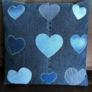 Peut inclure: Coussin carré en jean avec neuf patchs en forme de cœur dans différentes nuances de bleu. Le coussin a une fermeture éclair et est fabriqué à partir de denim recyclé. Les cœurs sont cousus sur le coussin avec du fil blanc, créant un motif patchwork.