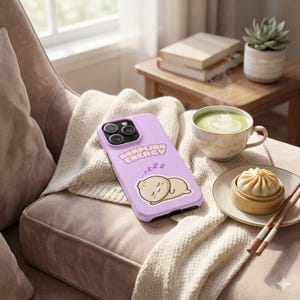 May include: A lavender phone case with the text "Dumpling Energy" and a cartoon dumpling illustration. The phone case is resting on a beige blanket on a chair. A cup of matcha and a dumpling are also visible.