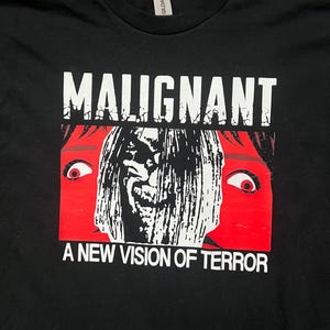 Puede incluir: Camiseta negra con un diseño gráfico. El diseño presenta la palabra "MALIGNANT" sobre una cara estilizada y aterradora con ojos rojos y el texto "A NEW VISION OF TERROR". El diseño es en blanco y rojo.