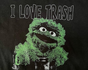 May include: Black t-shirt with the text "I LOVE TRASH" in white outlined letters. Below is a green and black graphic of a cartoon character with large eyes and a wide smile.