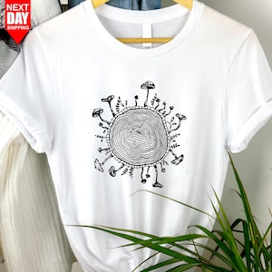 Könnte beinhalten: Weißes T-Shirt mit einer schwarz-weißen Illustration eines Baumstumpfes, umgeben von Pilzen und Blättern.