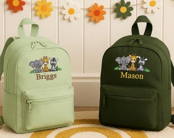 May include: Two small backpacks, one light green and one dark green, each with a front pocket and a top handle. The light green backpack has the name "Briggs" embroidered below a cartoon elephant, giraffe, and zebra. The dark green backpack has the name "Mason".