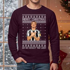 Puede incluir: Un jersey navide&ntilde;o granate con un dise&ntilde;o festivo. El jersey presenta el texto "MERRY CHRISTMAS" y "HO HO HOWAY THE LADS!" junto con un gr&aacute;fico de una persona. El dise&ntilde;o incluye copos de nieve y &aacute;rboles de Navidad.