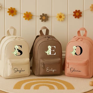 May include: Three personalized backpacks in cream, brown, and peach. Each has a letter and a dinosaur graphic. The cream backpack says "Skyler", the brown says "Evelyn", and the peach says "Olivia". A decorative garland of felt flowers hangs above.