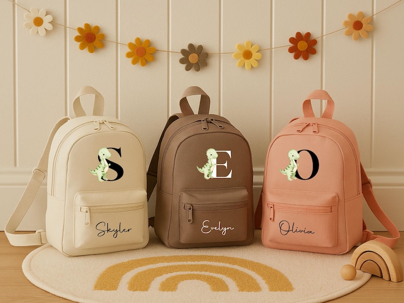 May include: Three personalized backpacks in cream, brown, and peach. Each has a letter and a dinosaur graphic. The cream backpack says "Skyler", the brown says "Evelyn", and the peach says "Olivia". A decorative garland of felt flowers hangs above.