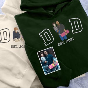May include: A green hoodie with a white "DAD" graphic and the text "EST. 2021". The graphic features a father and son holding a skateboard.