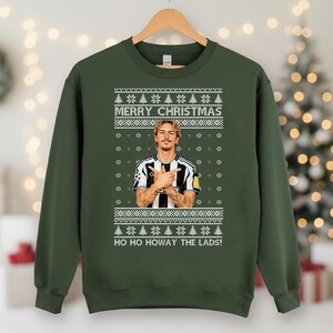 Puede incluir: Sudadera verde con un dise&ntilde;o festivo. La sudadera presenta un retrato de un hombre con los brazos cruzados, el texto "MERRY CHRISTMAS" y "HO HO HOWAY THE LADS!". El dise&ntilde;o incluye copos de nieve y &aacute;rboles de Navidad.