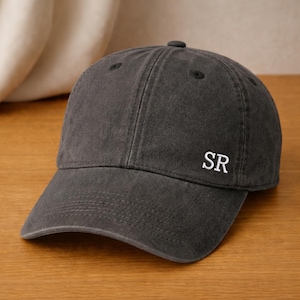 May include: A charcoal gray baseball cap with a curved brim. The cap features the white embroidered letters "SR" on the front. The hat is made of a soft, textured fabric and has a button on top.