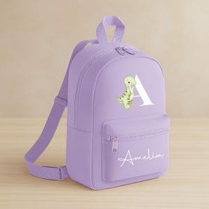 May include: A lavender backpack with a front pocket and adjustable straps. The backpack features a white letter "A" with a green dinosaur illustration and the name "Amelia" in white script. The bag has a top handle and a zipper closure.