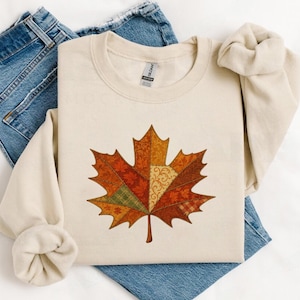 Op de afbeelding: Crèmekleurige sweatshirt met een groot, kleurrijk esdoornbladontwerp. Het blad bestaat uit verschillende stofpatronen in tinten oranje, bruin en groen. De sweatshirt wordt gecombineerd met een blauwe spijkerbroek.