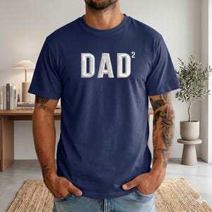 May include: Navy blue t-shirt with the word "DAD²" in a white, 3D-effect font. The shirt is a solid color and has short sleeves. The text is centered on the chest.