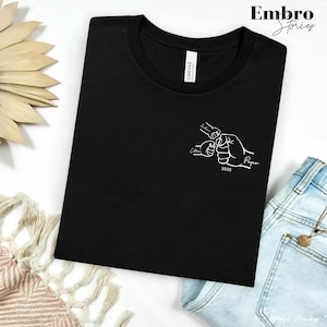 May include: A black short-sleeved t-shirt featuring a white line art graphic on the chest. The design depicts three fists bumping together with the names Jackson and Colton written above and beside them, the word Papa, and the year 2025.