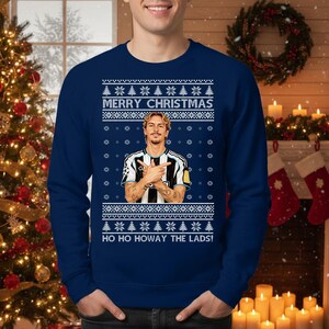 Puede incluir: Jersey navide&ntilde;o azul marino con un dise&ntilde;o festivo. El jersey presenta el texto "MERRY CHRISTMAS" y "HO HO HOWAY THE LADS!" junto con un gr&aacute;fico de una persona. El jersey tiene cuello redondo.