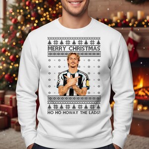 Puede incluir: Jersey navide&ntilde;o blanco con un dise&ntilde;o festivo. El jersey presenta el texto "MERRY CHRISTMAS" y "HO HO HOWAY THE LADS!". Tambi&eacute;n incluye un gr&aacute;fico de una persona con los brazos cruzados. El jersey tiene un patr&oacute;n de copos de nieve y &aacute;rboles de Navidad.