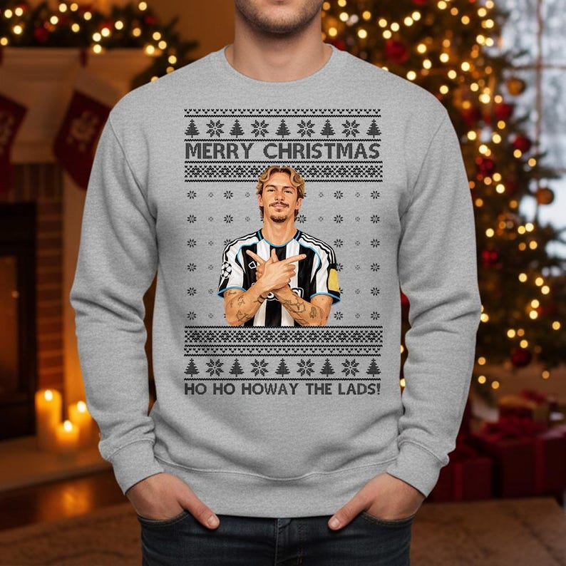 Puede incluir: Un jersey navide&ntilde;o gris con un dise&ntilde;o festivo. El jersey presenta el texto "MERRY CHRISTMAS" y "HO HO HOWAY THE LADS!". Tambi&eacute;n incluye un gr&aacute;fico de una persona con una camiseta de f&uacute;tbol. El jersey tiene cuello redondo y mangas largas.