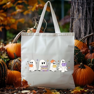 May include: White tote bag featuring a Halloween-themed design. The design includes four cartoon ghosts reading books, with stars and purple diamonds. Pumpkins and fall foliage are in the background.
