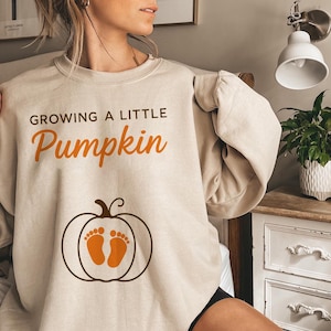 May include: Beige crew neck sweatshirt featuring the text "GROWING A LITTLE Pumpkin" in black and orange. The design includes a pumpkin graphic with baby footprints. A comfortable, casual top.