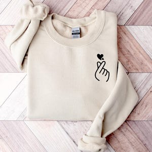 May include: A cream-coloured sweatshirt with a crew neck and long sleeves. A black heart and hand gesture graphic is printed on the front. The sweatshirt is folded on a light wood-grain surface.