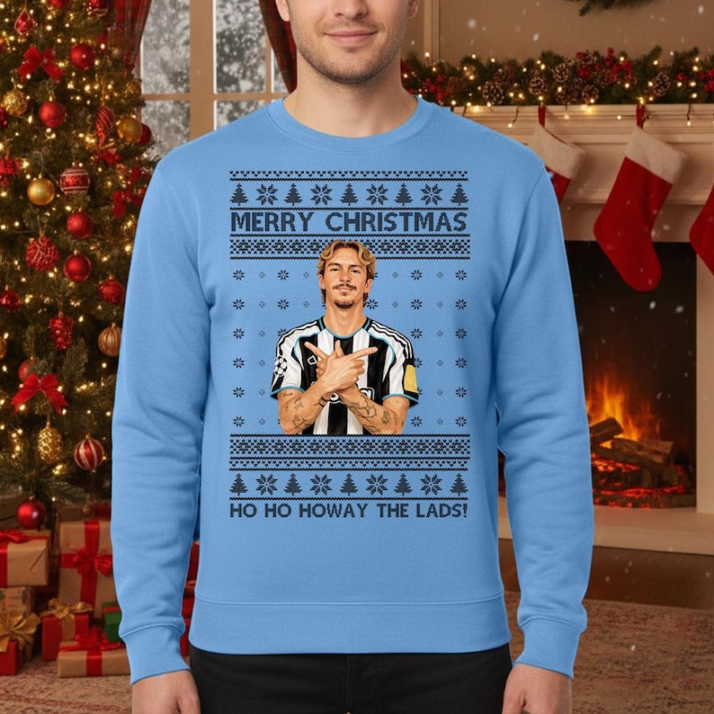 Puede incluir: Su&eacute;ter navide&ntilde;o azul claro con un dise&ntilde;o festivo. El su&eacute;ter presenta el texto "MERRY CHRISTMAS" y "HO HO HOWAY THE LADS!" junto con un gr&aacute;fico de una persona con una camiseta a rayas blancas y negras. El dise&ntilde;o incluye copos de nieve y &aacute;rboles de Navidad.
