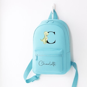 May include: A light blue backpack with a front pocket and adjustable straps. The backpack features a dinosaur illustration next to the letter "C" and the name "Charlotte" in script.