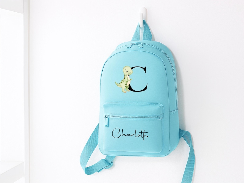 May include: A light blue backpack with a front pocket and adjustable straps. The backpack features a dinosaur illustration next to the letter "C" and the name "Charlotte" in script.