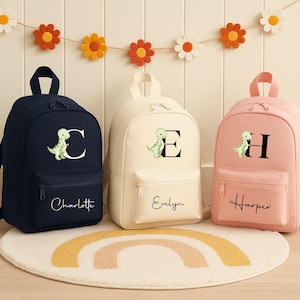 May include: Three personalized backpacks in navy blue, cream, and pink. Each features a dinosaur graphic, a large initial, and a name. The backpacks have a front pocket and adjustable straps. A decorative garland of felt flowers hangs above.