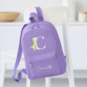 May include: A lilac backpack with a front zippered pocket and adjustable straps. The backpack features a green and yellow dinosaur illustration with the letter "C" and the name "Charlotte" in white script.