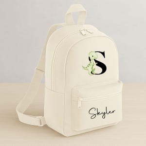 May include: Cream-colored backpack with a front pocket and adjustable straps. The backpack features a green dinosaur graphic next to a black letter "S" and the name "Skyler" in cursive.
