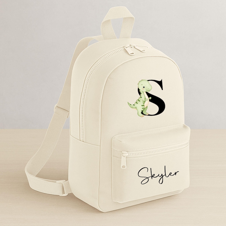 May include: Cream-colored backpack with a front pocket and adjustable straps. The backpack features a green dinosaur graphic next to a black letter "S" and the name "Skyler" in cursive.