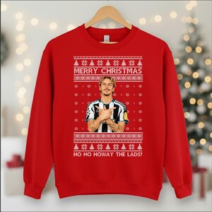 Puede incluir: Una sudadera roja con un dise&ntilde;o festivo. La sudadera presenta el texto "MERRY CHRISTMAS" encima de una imagen de una persona, y la frase "HO HO HOWAY THE LADS!" debajo. El dise&ntilde;o incluye copos de nieve y &aacute;rboles de Navidad.