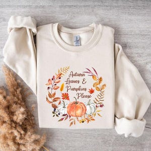 May include: Cream-coloured sweatshirt with rolled sleeves. The front features a watercolour wreath with the text "Autumn Leaves & Pumpkins Please." The wreath includes a pumpkin, white pumpkins, and autumn foliage.